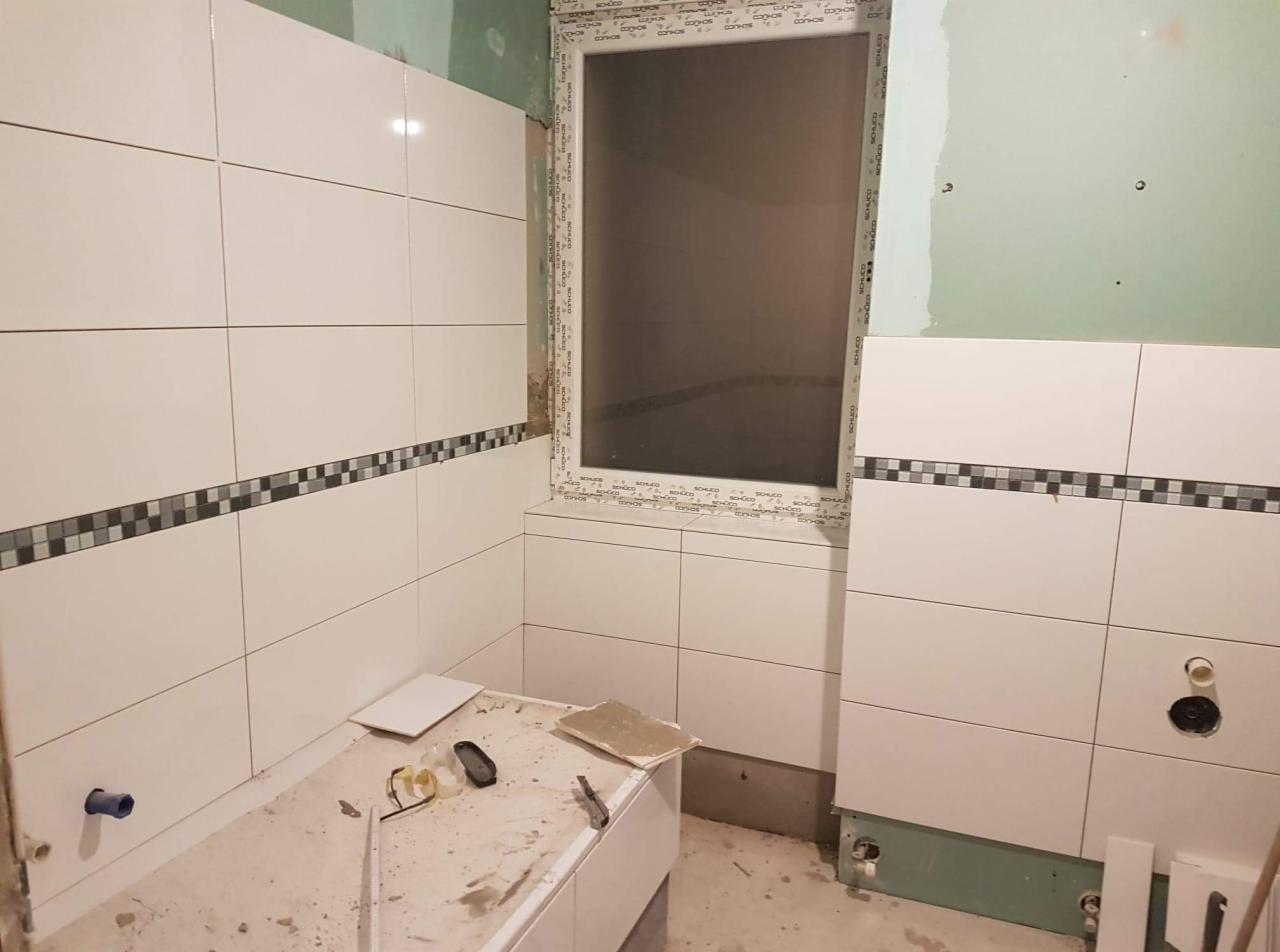 Białe płytki ścienne z dekoracyjnym pasem mozaiki w łazience w trakcie remontu, widoczne narzędzia i niezakończone prace przy oknie.