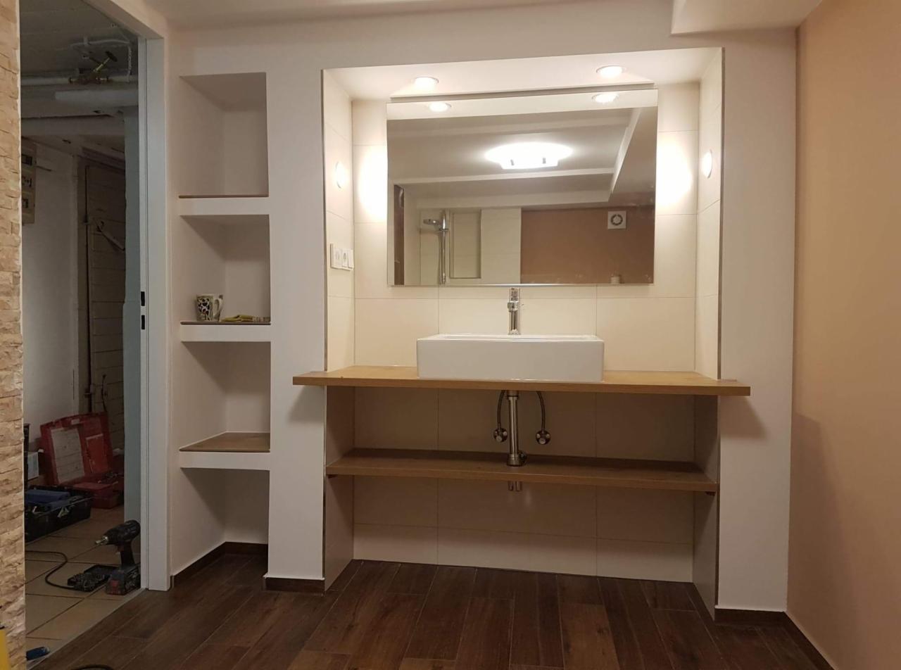 Wykończona łazienka z umywalką nablatową, lustrem z oświetleniem, drewnianym blatem i półkami w zabudowie, widoczna wiertarka i narzędzia w tle.