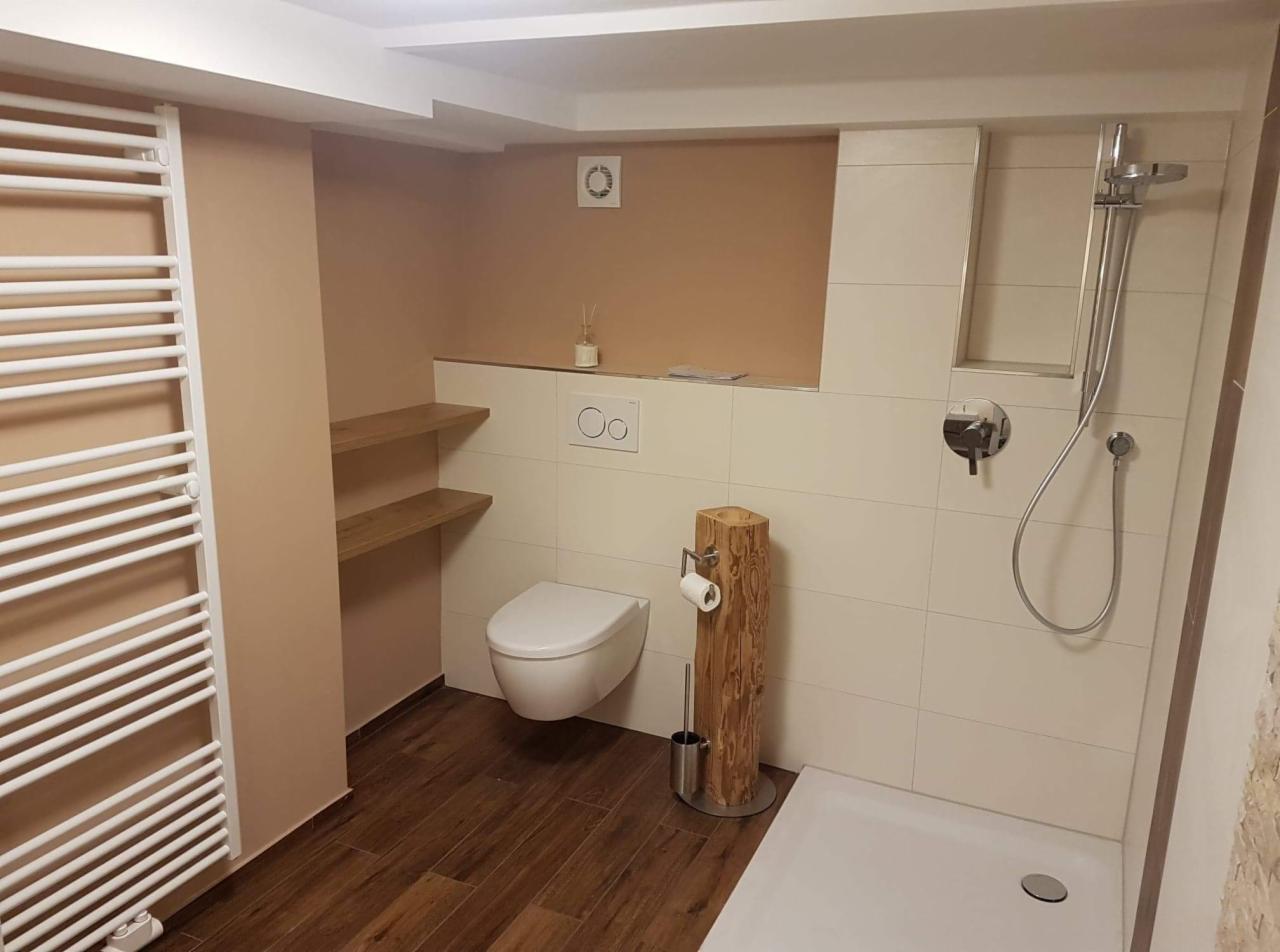 Nowoczesna łazienka z brązowymi akcentami: drewniana podłoga, półki i uchwyt na papier toaletowy z pnia, biała armatura i grzejnik drabinkowy.