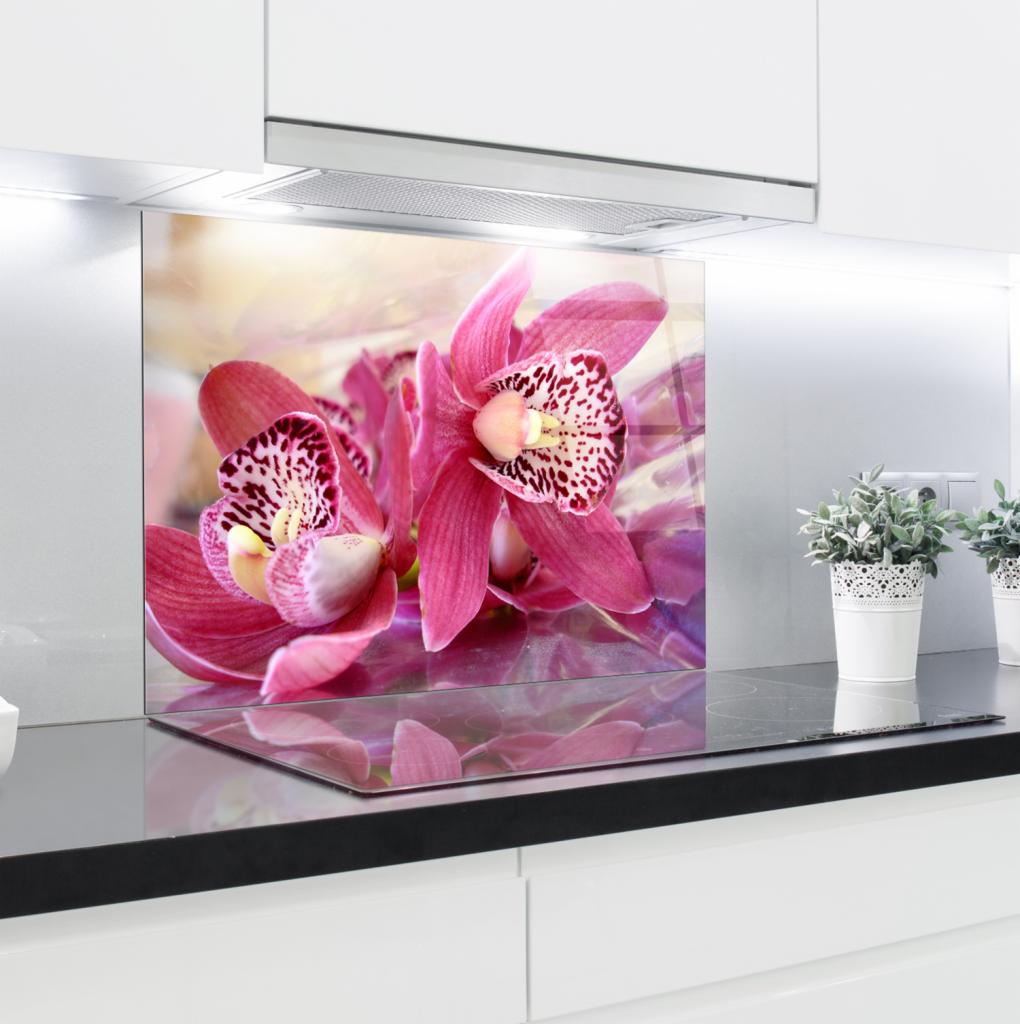 Szklany panel kuchenny z motywem różowych orchidei, zamontowany nad płytą indukcyjną, w tle białe szafki i okap.