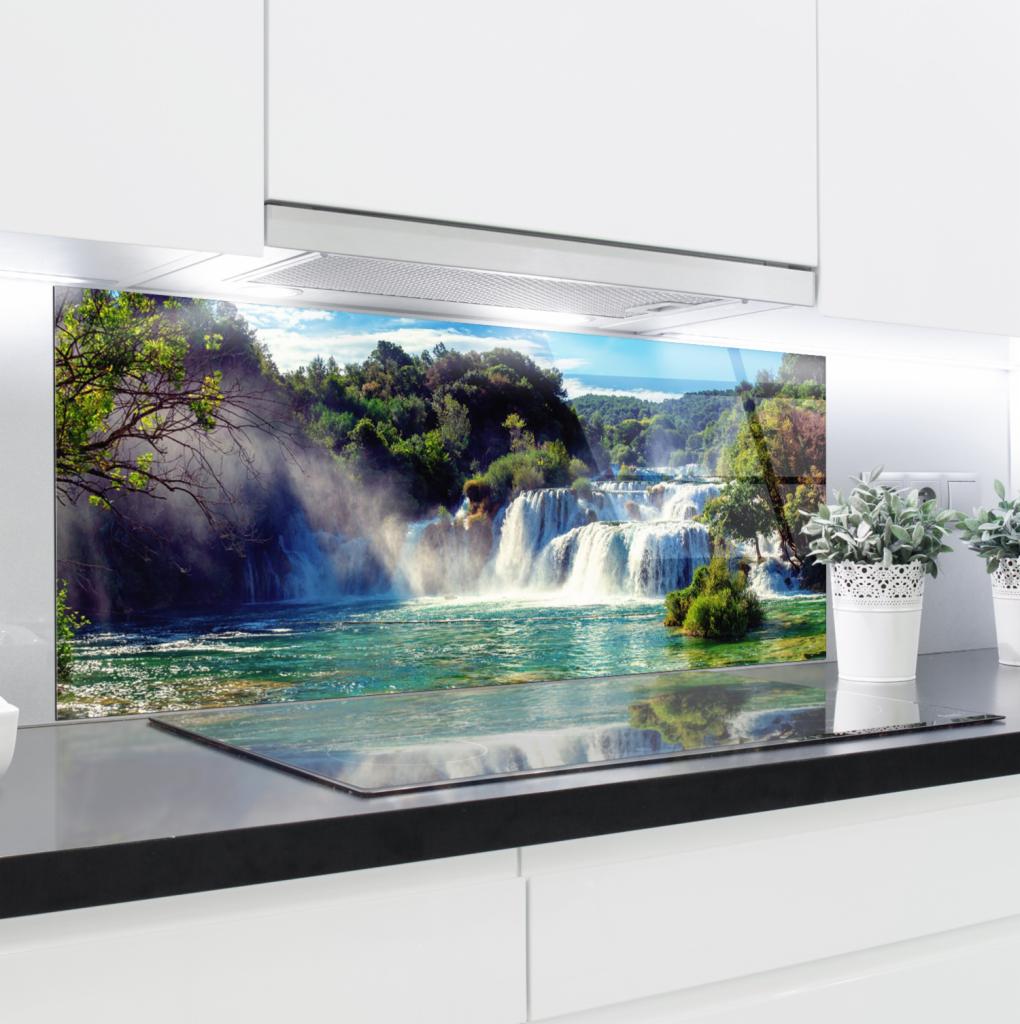 Szklany panel kuchenny z nadrukiem wodospadu, zamontowany między blatem a szafkami, odbijający się w płycie grzewczej, z dwoma doniczkami ziół po prawej stronie.