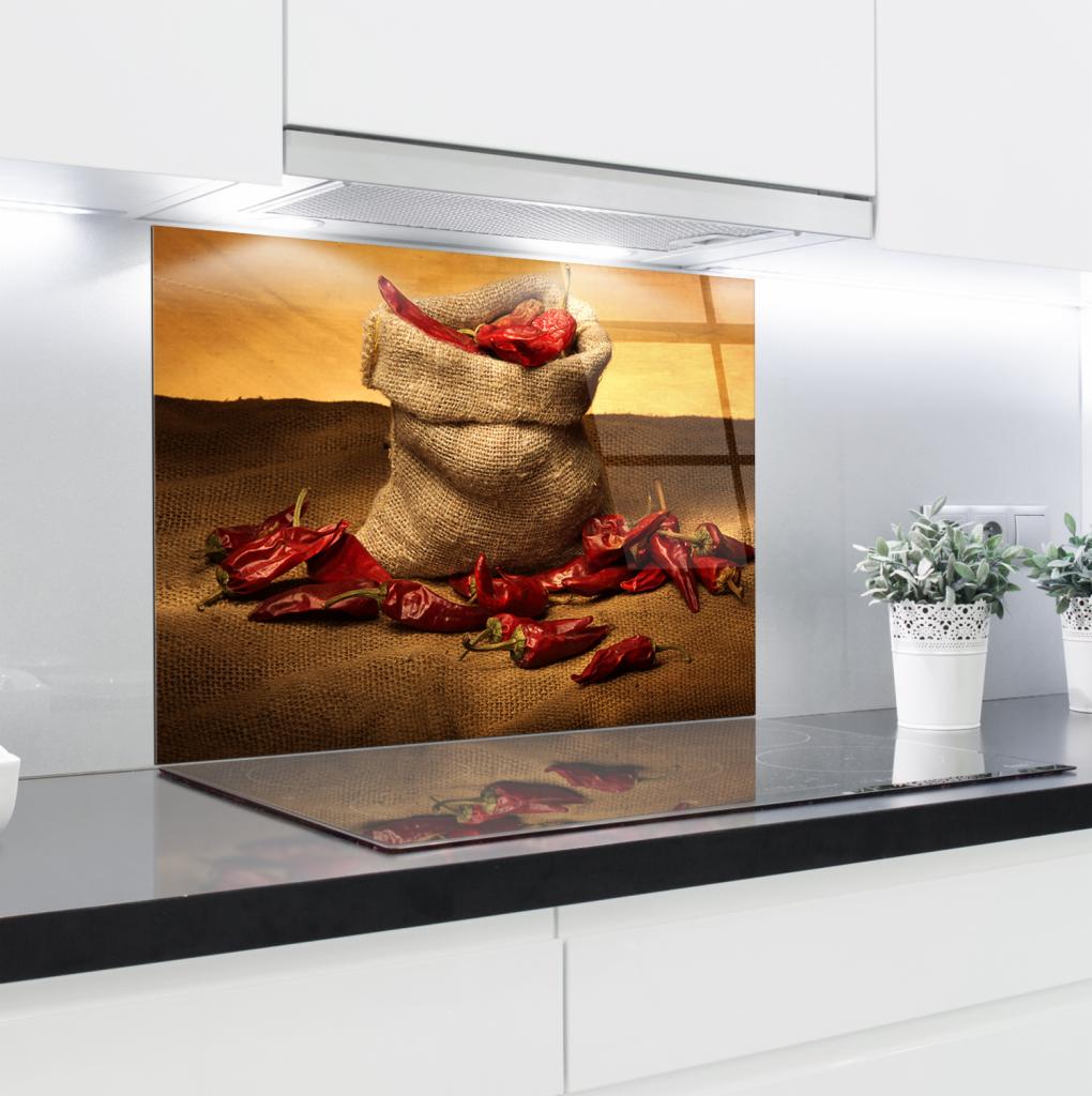 Szklany panel kuchenny z motywem worka z papryczkami chili, odbijający się w płycie indukcyjnej, z widocznymi doniczkami z ziołami w tle.