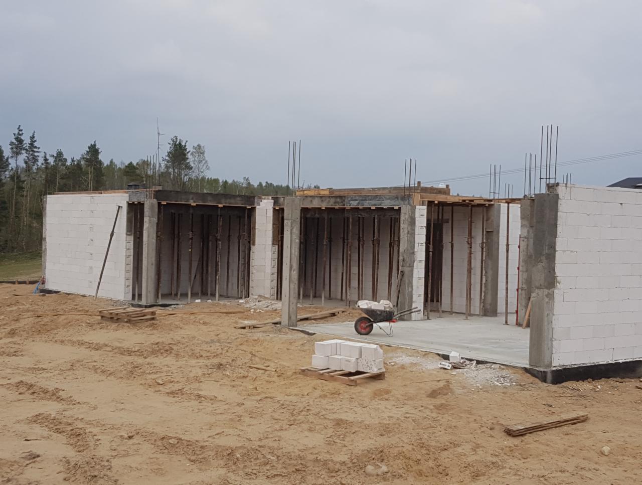 Wznoszenie ścian z bloczków betonu komórkowego na fundamencie, widoczne zbrojenia pionowe i szalunki pod wieniec, taczka budowlana obok