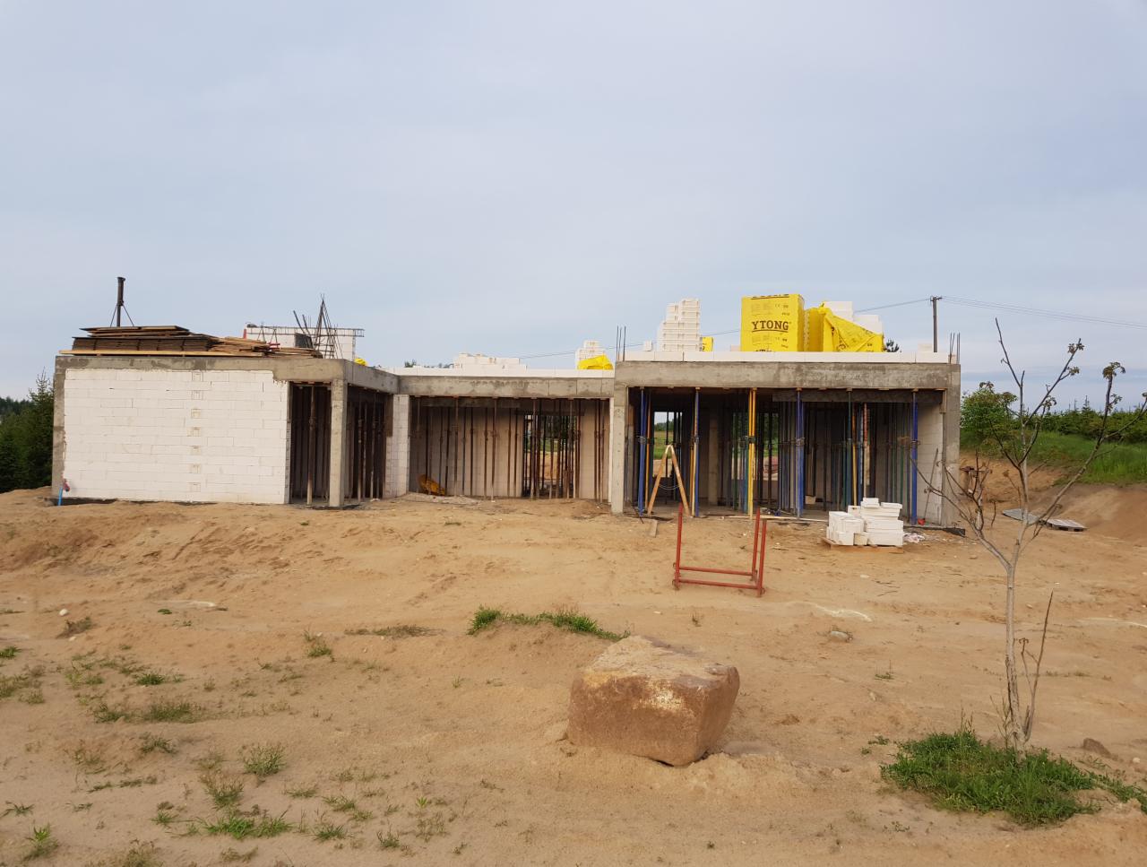 Surowy stan domu z betonu komórkowego Ytong w trakcie budowy, widoczne podpory szalunkowe, teren budowy z piaskiem i kamieniem.