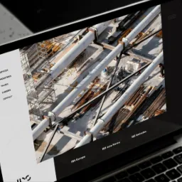 Laptop wyświetlający stronę internetową firmy IBS z widokiem z góry na plac budowy z rurami i rusztowaniami.