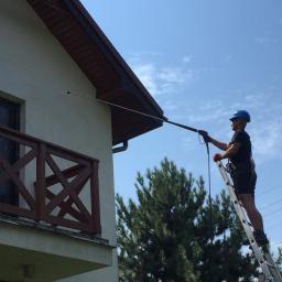 RBN Polska Sp. z o.o. - Mężczyzna w kasku i uprzęży na drabinie czyści elewację domu za pomocą myjki ciśnieniowej z długą lancą, w tle błękitne niebo i zieleń drzew.