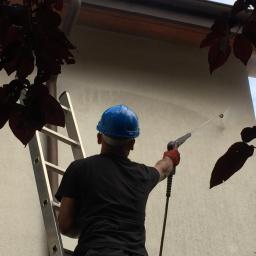 RBN Polska Sp. z o.o. - Pracownik w niebieskim kasku, na drabinie, myje elewację budynku myjką ciśnieniową, widoczne liście drzew