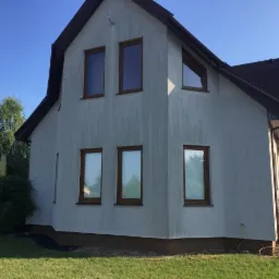 Dwupiętrowy dom z szarą elewacją i brązowymi ramami okiennymi, otoczony zielonym trawnikiem i drzewami w słoneczny dzień.