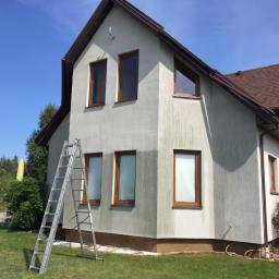 RBN Polska Sp. z o.o. - Dwupiętrowy dom z szarą elewacją w trakcie czyszczenia, widoczna różnica w kolorze ściany, aluminiowa drabina oparta o ścianę, brązowe ramy okienne, zielony trawnik i drzewa w tle.