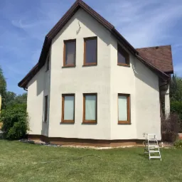 Elewacja domu jednorodzinnego po remoncie, jasny tynk, brązowe okna i dachówka, drabina oparta o ścianę, zielony trawnik.