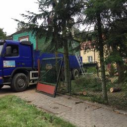 RBN Polska Sp. z o.o. - Wycinka drzew: niebieska ciężarówka z podnośnikiem i drabiną na tle domu, obok pnia ściętych drzew i otwartej bramy.