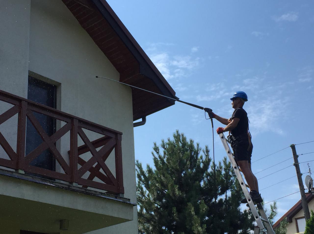 Mężczyzna w kasku i uprzęży na drabinie czyści elewację domu za pomocą myjki ciśnieniowej z długą lancą, w tle błękitne niebo i zieleń drzew.