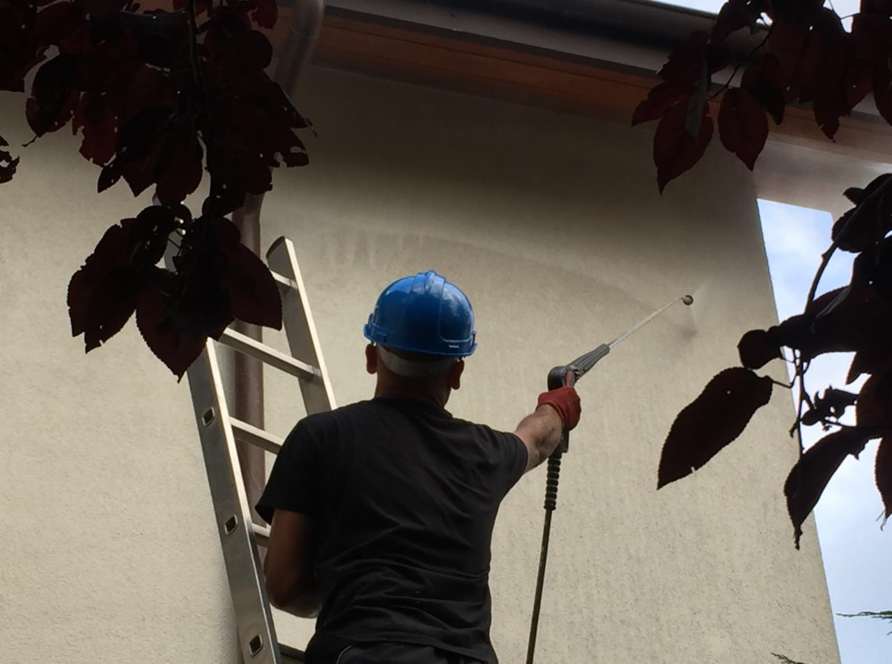 Pracownik w niebieskim kasku, na drabinie, myje elewację budynku myjką ciśnieniową, widoczne liście drzew