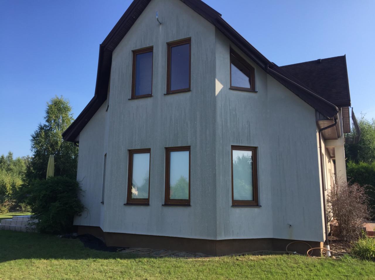 Dwupiętrowy dom z szarą elewacją i brązowymi ramami okiennymi, otoczony zielonym trawnikiem i drzewami w słoneczny dzień.