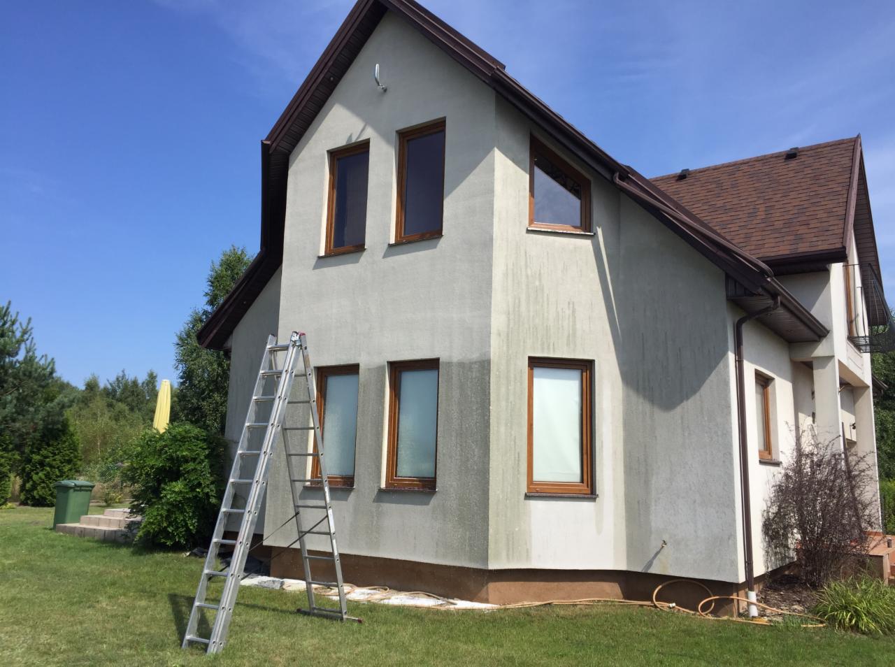 Dwupiętrowy dom z szarą elewacją w trakcie czyszczenia, widoczna różnica w kolorze ściany, aluminiowa drabina oparta o ścianę, brązowe ramy okienne, zielony trawnik i drzewa w tle.