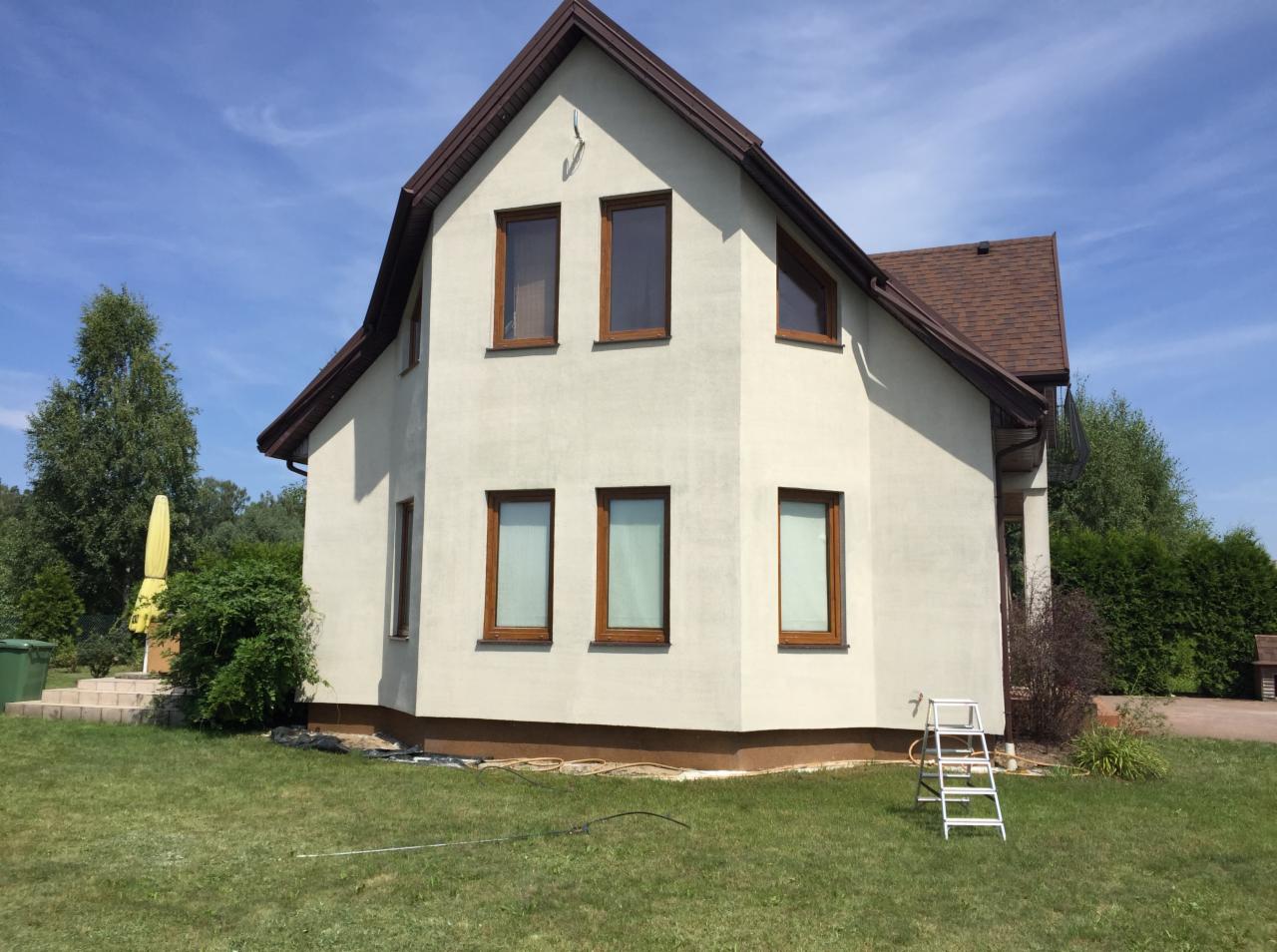 Elewacja domu jednorodzinnego po remoncie, jasny tynk, brązowe okna i dachówka, drabina oparta o ścianę, zielony trawnik.