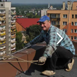 Dekarz w niebieskiej czapce i wzorzystej koszuli klęczy na dachu, trzymając linę, z widokiem na miasto w tle.