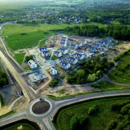 Widok z lotu ptaka na osiedle domów jednorodzinnych w trakcie budowy, z widocznym rondem i otaczającą zielenią.
