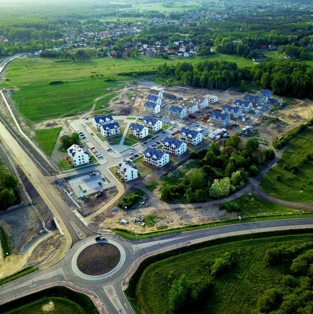 Widok z lotu ptaka na osiedle domów jednorodzinnych w trakcie budowy, z widocznym rondem i otaczającą zielenią.