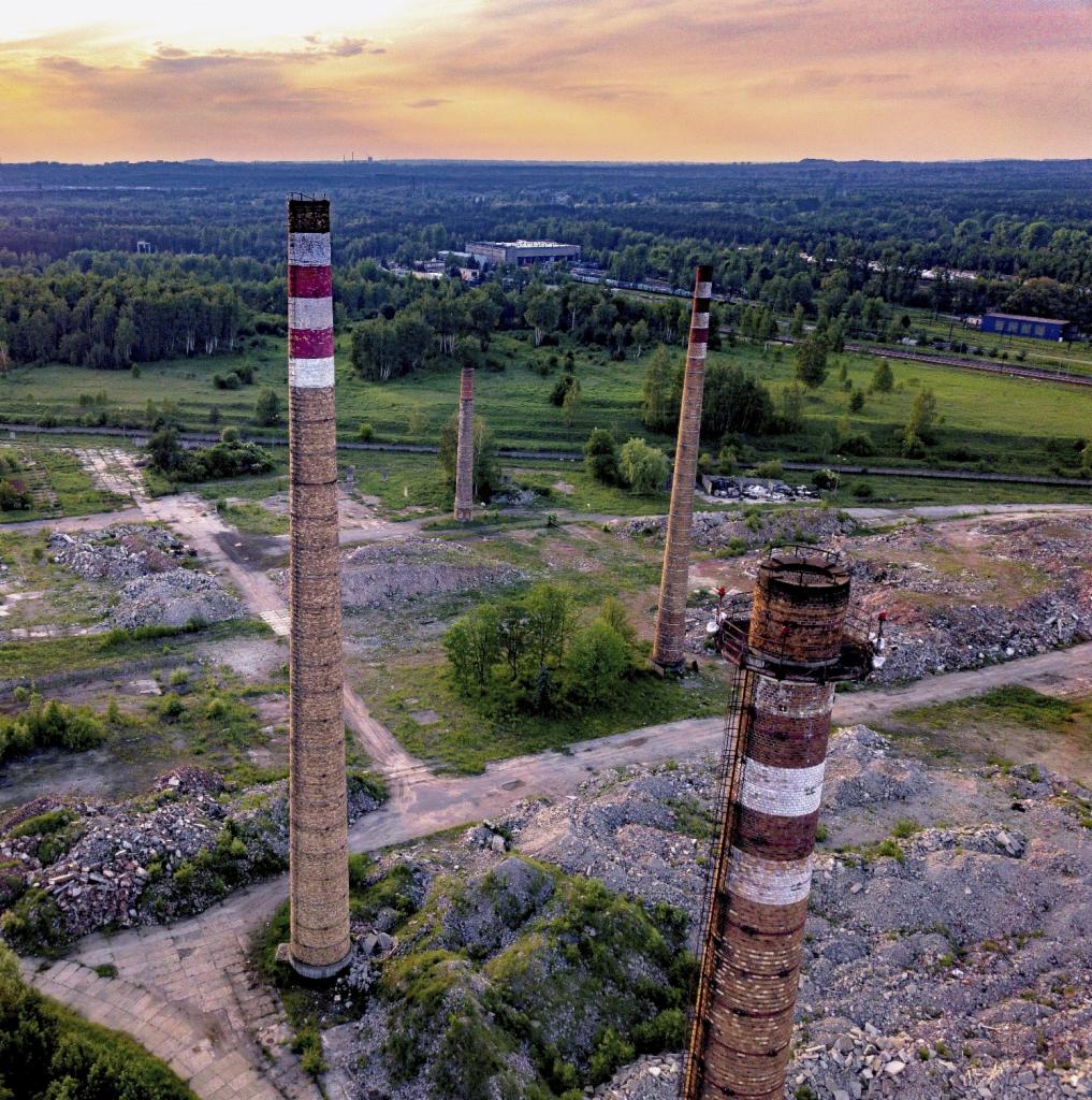 Widok z drona na teren poprzemysłowy z trzema ceglanymi kominami w różnym stanie zachowania, otoczony zielenią i lasami, pod wieczornym niebem.