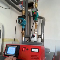 CPS SERVICE Waldemar Dobosz - Czerwona obudowa urządzenia z wyświetlaczem i przyciskami, obok instalacja rur z zaworami i pompami obiegowymi, widoczne elementy instalacji grzewczej w pomieszczeniu.