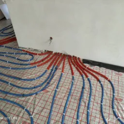 Rozłożone rury ogrzewania podłogowego, czerwone i niebieskie, na folii z nadrukiem siatki, w surowym wnętrzu przed wylaniem posadzki.