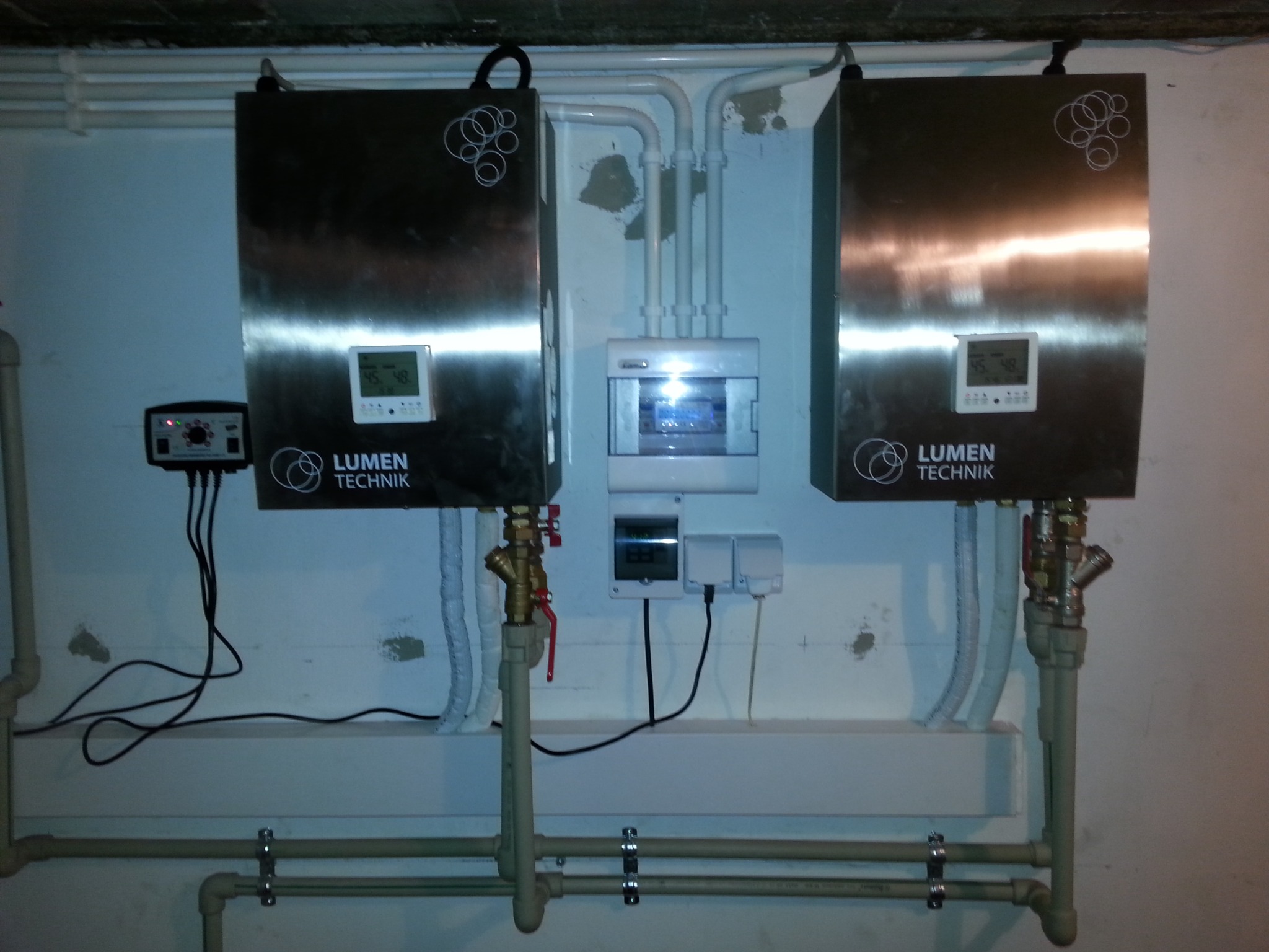 Dwa metalowe urządzenia grzewcze marki Lumen Technik zamontowane na ścianie, z widocznymi rurami, zaworami i przewodami elektrycznymi, oraz skrzynką rozdzielczą.