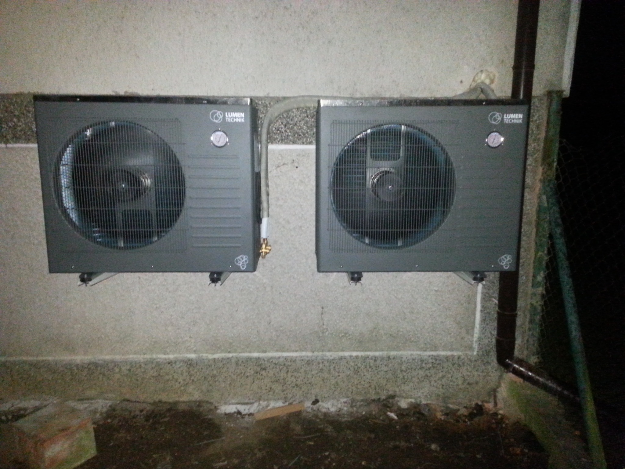 Dwa szare urządzenia Lumen Technik zamontowane na ścianie budynku, połączone rurą. Widoczne elementy instalacji i fragment rynny. Niska jakość oświetlenia.