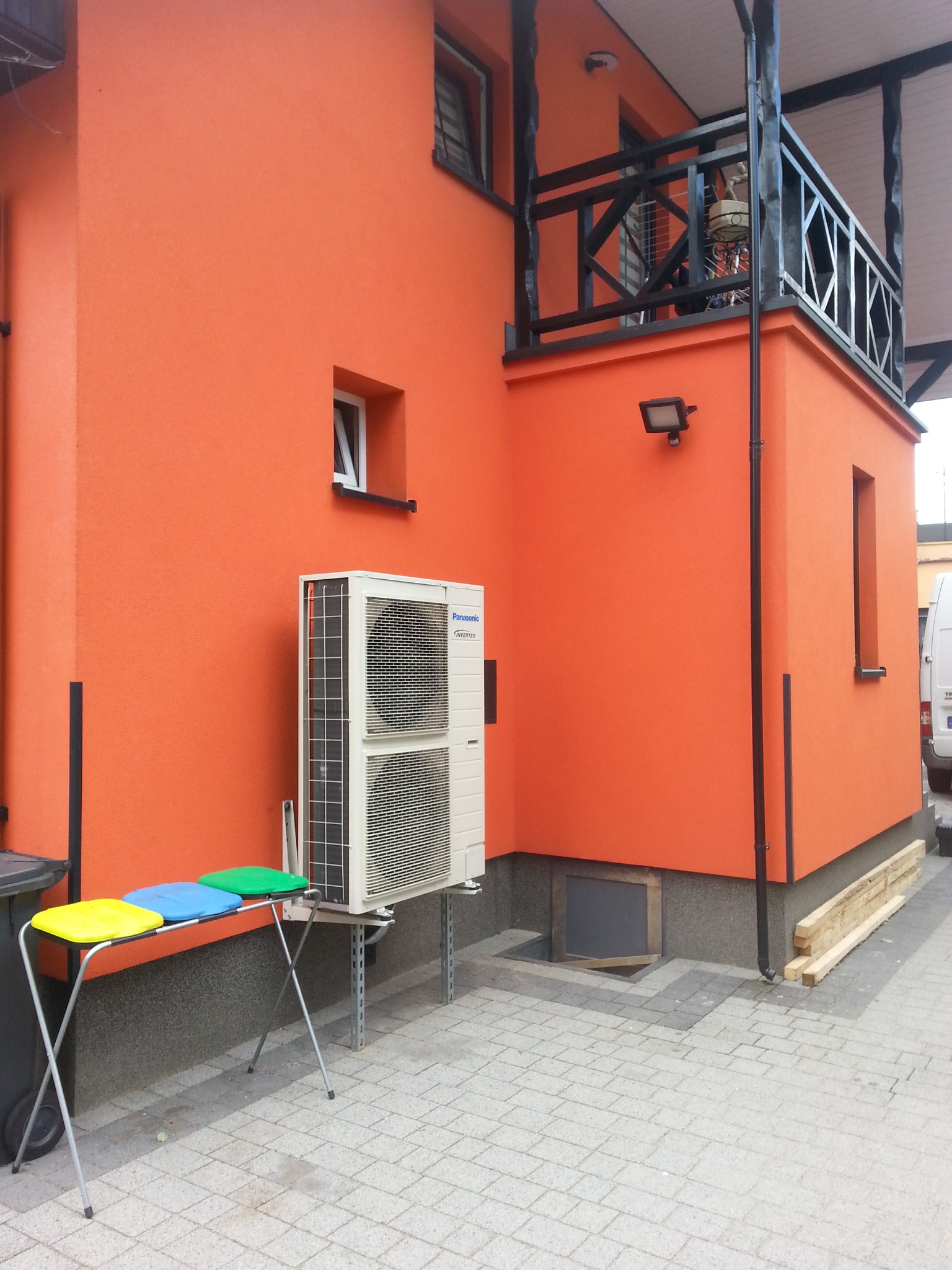 Pompa ciepła Panasonic Inverter zamontowana na zewnątrz budynku o pomarańczowej elewacji, widoczny fragment balkonu z czarną balustradą oraz trzy kolorowe siedziska na składanym stole.