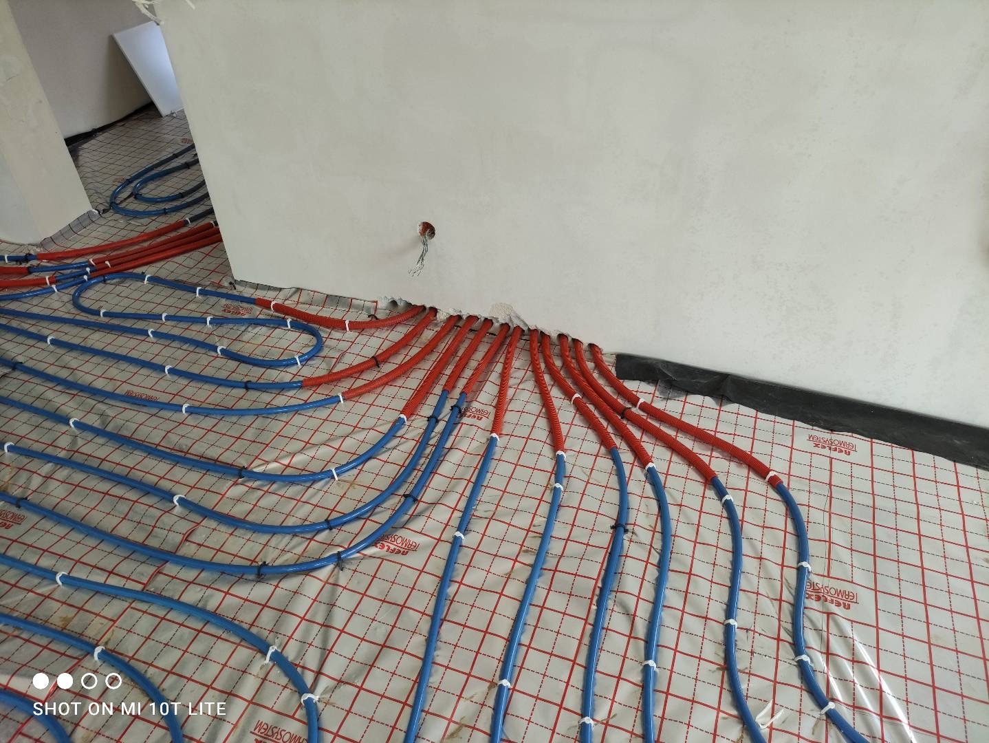 Rozłożone rury ogrzewania podłogowego, czerwone i niebieskie, na folii z nadrukiem siatki, w surowym wnętrzu przed wylaniem posadzki.