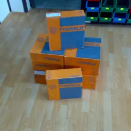 Stos pudełek z drutem spawalniczym Tysweld 1,2mm na podłodze w Rybniku, widoczne regały z pojemnikami w tle.