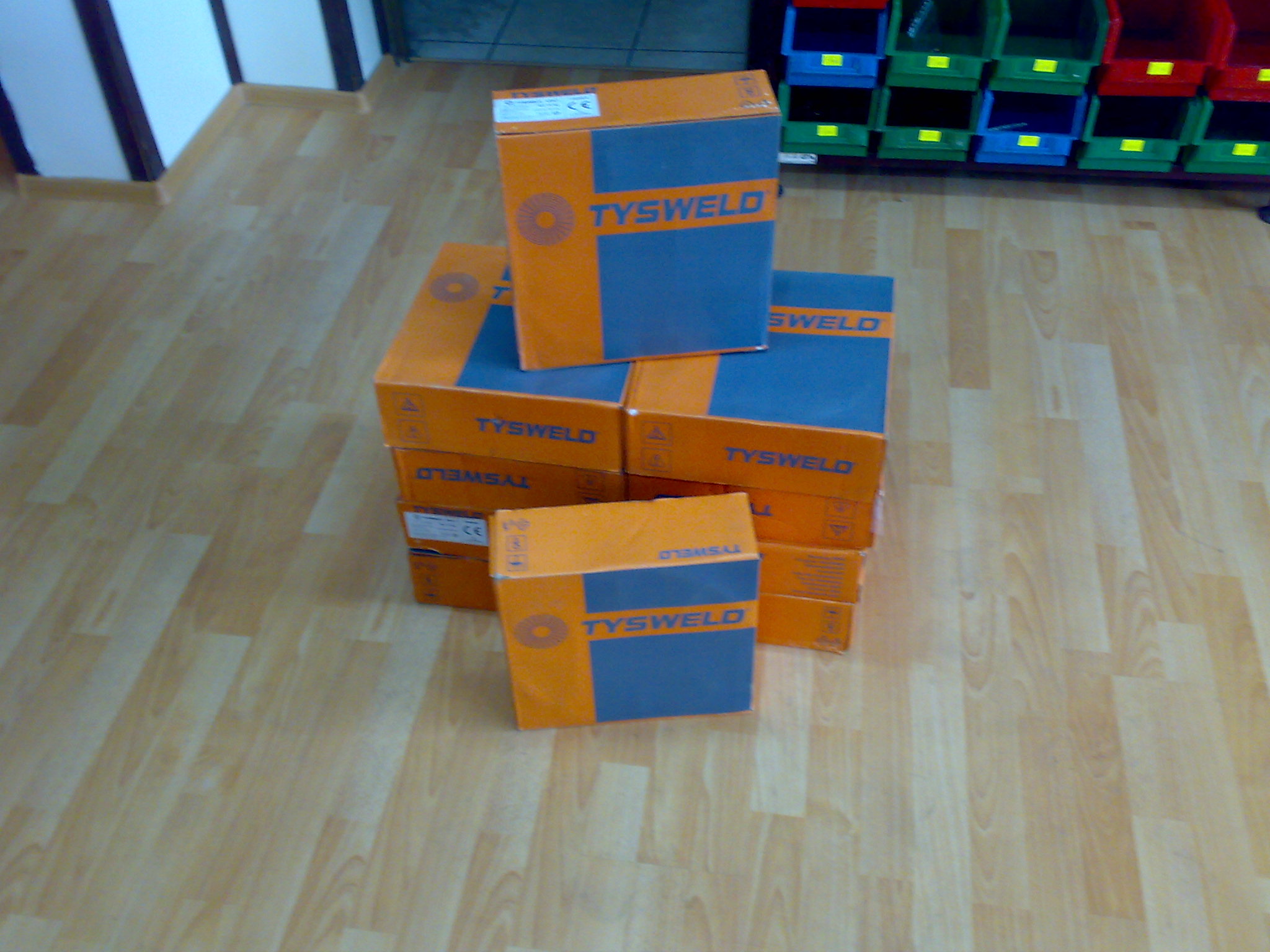 Stos pudełek z drutem spawalniczym Tysweld 1,2mm na podłodze w Rybniku, widoczne regały z pojemnikami w tle.