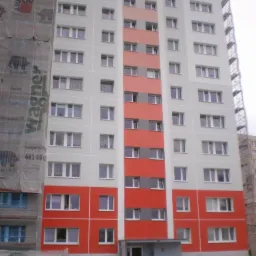 Wielopiętrowy blok mieszkalny po renowacji elewacji z fragmentami w kolorze szarym i czerwonym, na części budynku widoczne rusztowania i reklama farb Wagner.