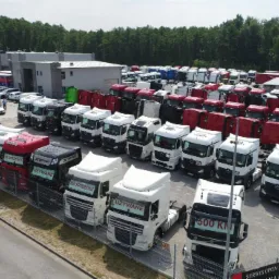 Plac z dużą ilością używanych ciągników siodłowych różnych marek i kolorów, z widocznymi tabliczkami 'Na sprzedaż' i 'Używane', na tle budynku firmy i drzew.