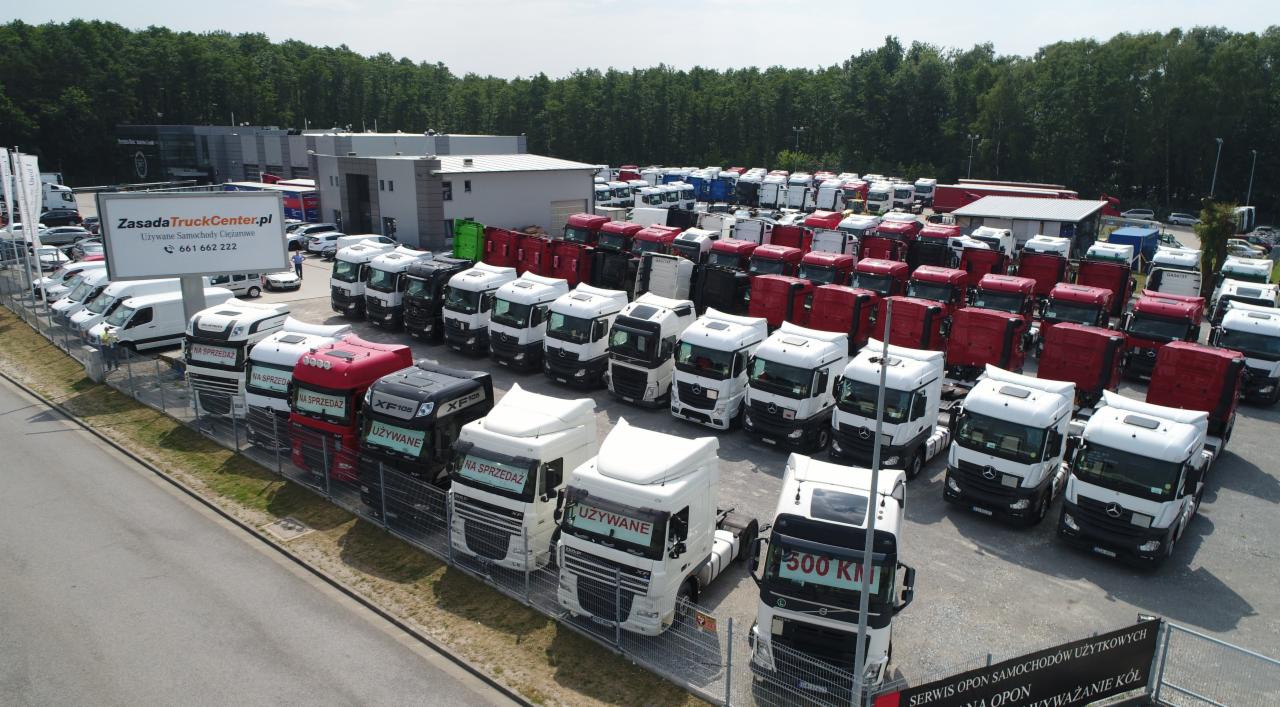 Plac z dużą ilością używanych ciągników siodłowych różnych marek i kolorów, z widocznymi tabliczkami 'Na sprzedaż' i 'Używane', na tle budynku firmy i drzew.