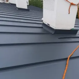 Szary blaszany dach z kilkoma białymi kominami, jeden z szarą rurą wentylacyjną i pomarańczową liną zabezpieczającą.