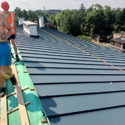 Ekipa dekarzy w trakcie montażu blachy na dachu, widoczne drewniane łaty i zielona membrana dachowa, w tle kominy i zieleń drzew.