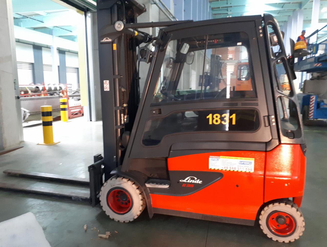 Pomarańczowy wózek widłowy Linde E35 z numerem 1831 na tle magazynu z otwartą bramą, widoczne żółto-czarne słupki ostrzegawcze oraz podnośnik koszowy w tle.