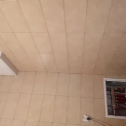 Beżowe płytki ceramiczne na ścianie i suficie łazienki, widoczna klapa rewizyjna z instalacją hydrauliczną i gniazdko elektryczne