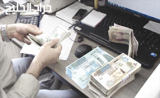 Na biurku leżą sterty banknotów w różnych walutach (dolary i dinary jordańskie), obok kalkulator, laptop i telefon komórkowy; dłoń liczy dolary.