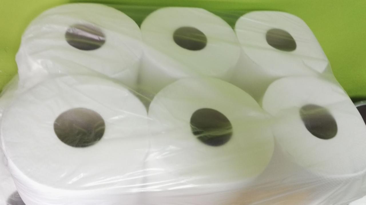 Sześć rolek białego papieru toaletowego, zapakowanych w przezroczysty plastik, ustawionych w dwóch rzędach na tle zielonej ściany.