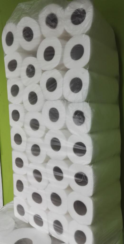 Duża paczka rolek białego papieru toaletowego, zapakowana w folię, ułożona w stos na tle zielonej ściany.