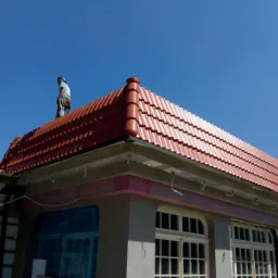 Dekarz na dachu z nową, czerwoną dachówką ceramiczną, widoczne okna budynku pod dachem.