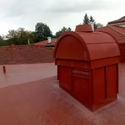 Czerwona, metalowa obudowa wentylacyjna na płaskim, czerwonym dachu pokrytym powłoką hydroizolacyjną, z widocznymi dachówkami w tle.