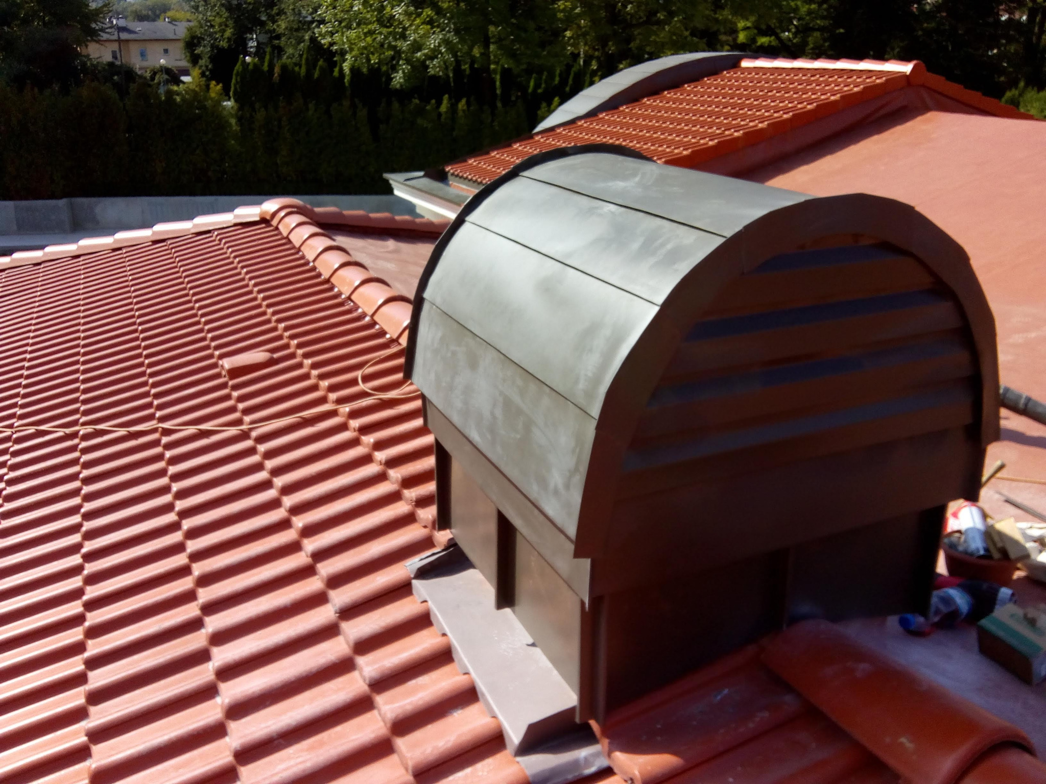 Nowa, ceramiczna dachówka w kolorze ceglastym z zamontowanym, metalowym wywietrznikiem dachowym z elementami wykończeniowymi, widok z góry.
