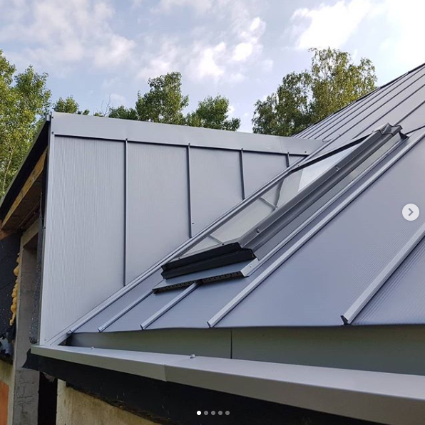 Szary dach z blachy na ukończonym budynku z oknem dachowym, widok z dołu pod kątem, na tle zielonych drzew i błękitnego nieba.