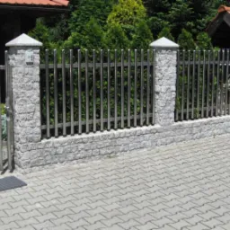 Nowoczesne ogrodzenie z metalowymi prętami i kamiennymi słupkami, z bramką wejściową i wycieraczką przed nią, na tle zadbanego ogrodu.
