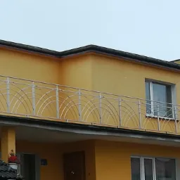 Żółty dom z balkonem zabezpieczonym metalową balustradą o wzorze łuków, widoczne fragmenty dachu sąsiedniego budynku.