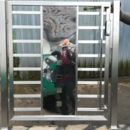 Nowoczesna brama wjazdowa ze stali nierdzewnej z lustrzanym panelem centralnym i elektronicznym zamkiem szyfrowym, widoczna osoba w stroju roboczym podczas montażu.