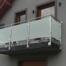 Balkon z matową szklaną balustradą i nierdzewnymi słupkami, widok z dołu na tle szarej elewacji domu z dwoma oknami.